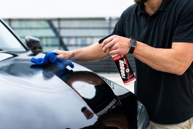 A person in a black polo shirt wipes the hood of a black car with a blue microfiber cloth while holding a spray bottle.