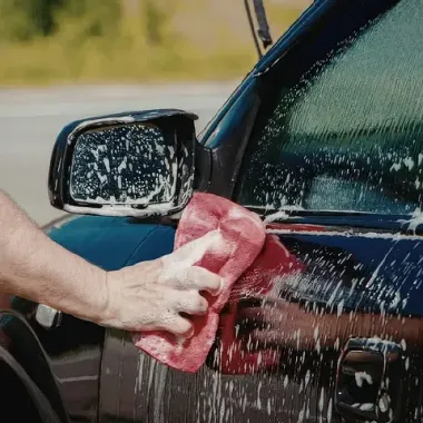 A hand holding a red sponge washes a dark car with soap suds near the side mirror.