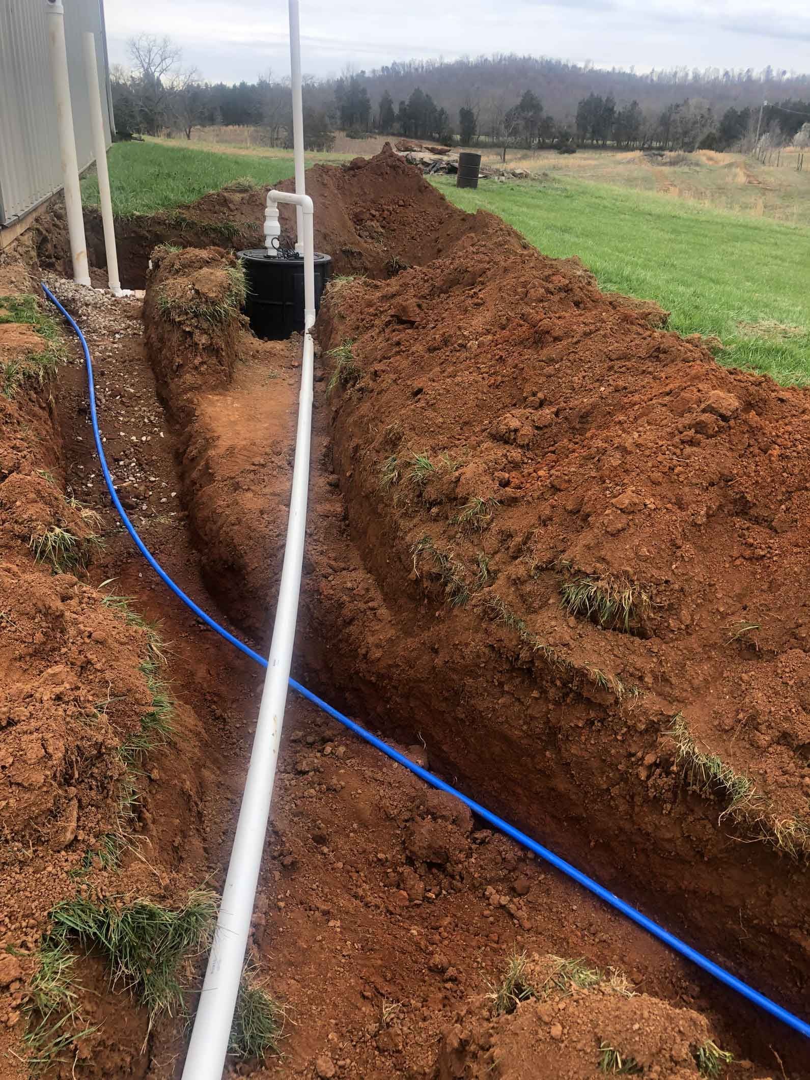 Trench dug in brown earth with white and blue pipes, near a building and field.