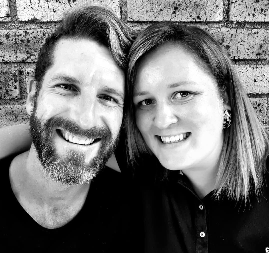 A black-and-white close-up portrait of two smiling people leaning their heads together in front of a brick wall.