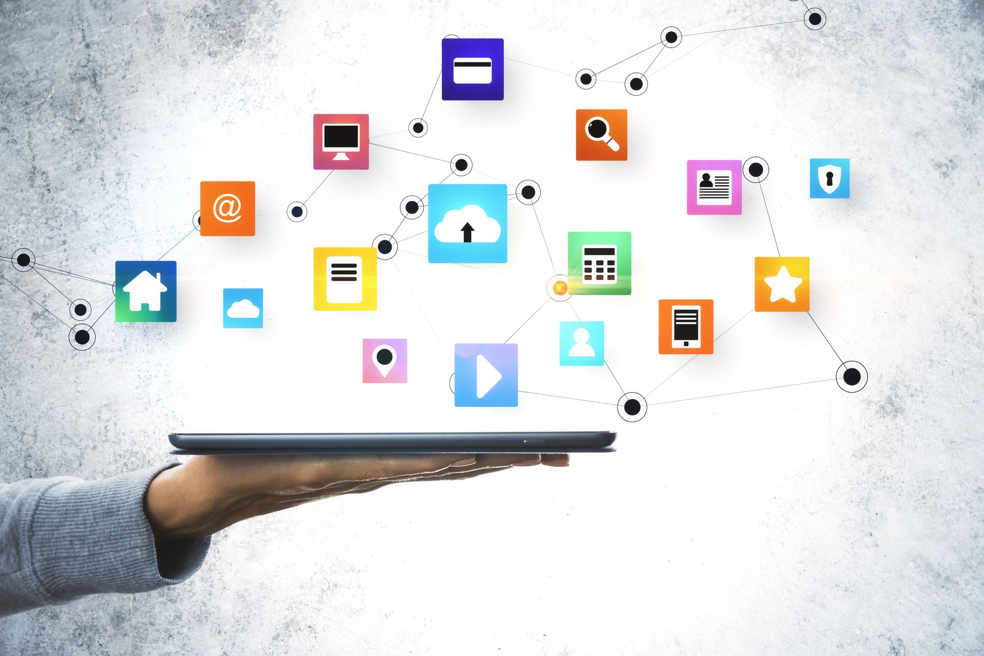 A hand holds a tablet displaying various colorful app icons connected by lines against a concrete background.