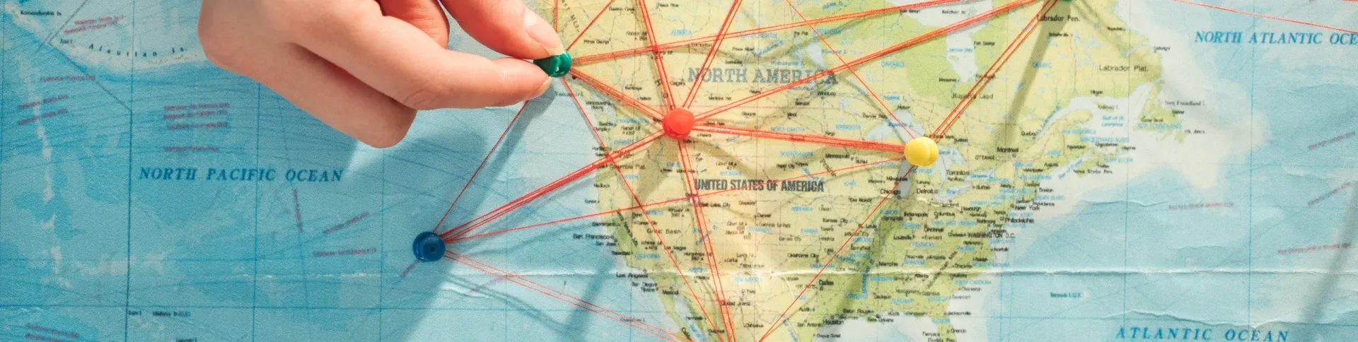 A hand places a green pin on a map of North America, where other colorful pins are connected by red string lines.