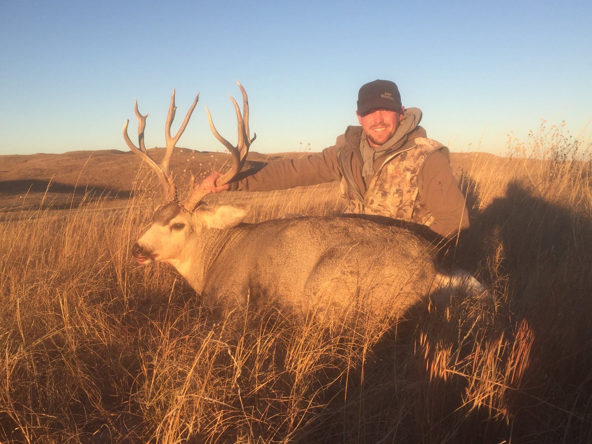 Man in camo vest poses with a large deer buck in a field at sunset.
