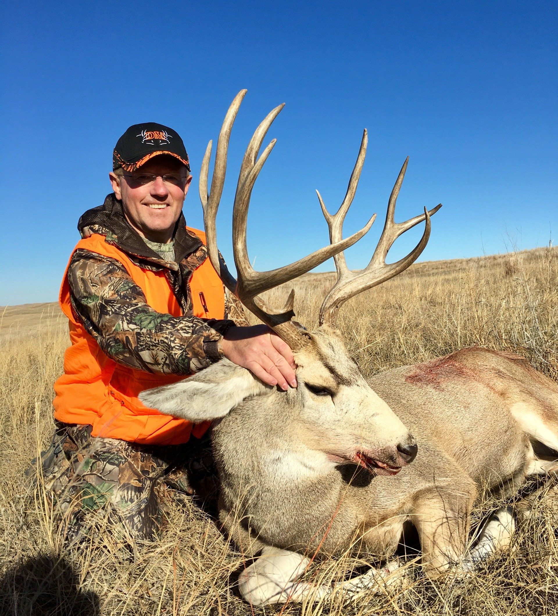 Hunter kneels next to a large mule deer buck. Man smiles wearing orange vest, deer on dry grass, blue sky.