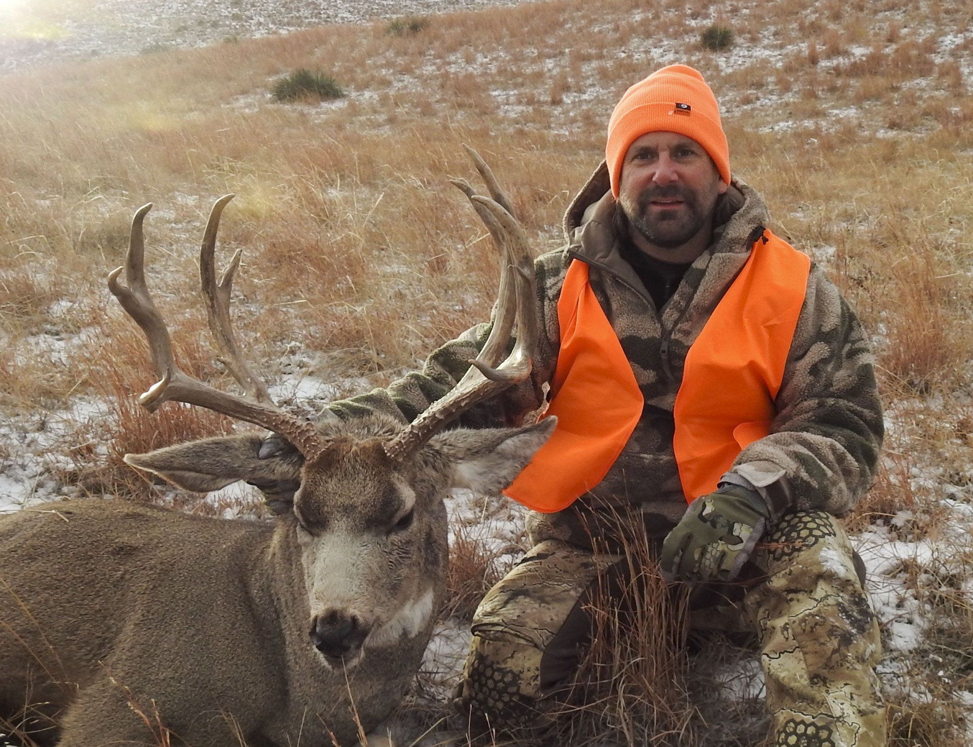 Man in orange vest kneels next to a large deer with antlers on a snow-covered hillside.