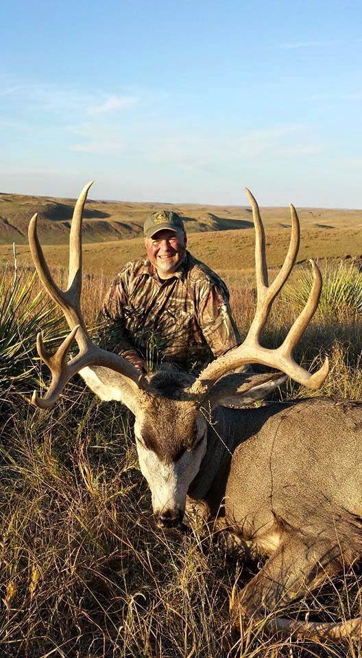 Man in camo with large buck, outdoors. Sunny field, blue sky. Man smiles, deer has large antlers.