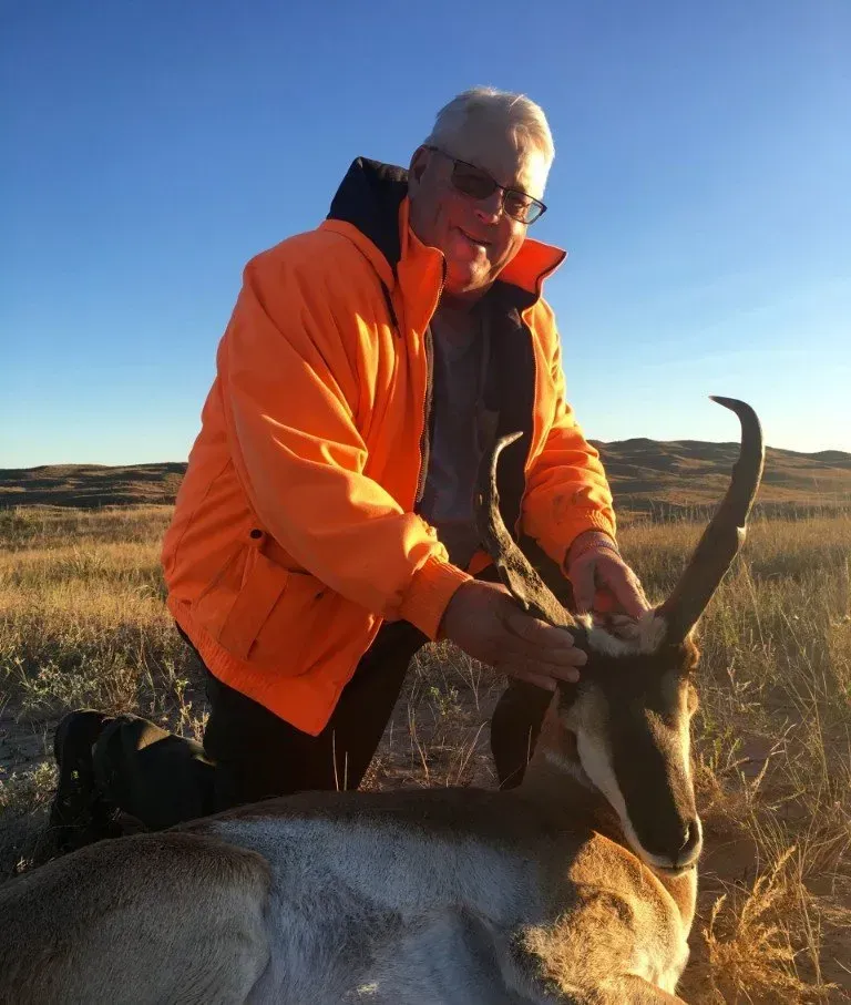 Man kneels in field next to a pronghorn antelope, wearing orange jacket, smiling.