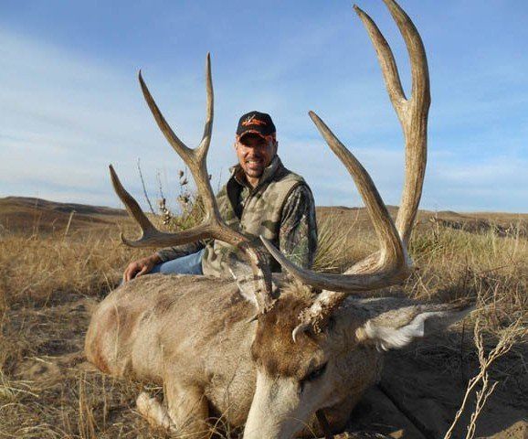 Man in camo kneels beside a large deer with large antlers, in a grassy field.