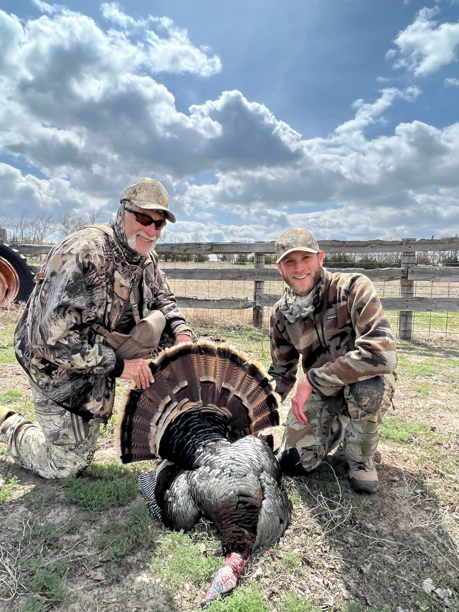 Two hunters in camouflage kneel beside a harvested turkey in a field; blue sky, white clouds.