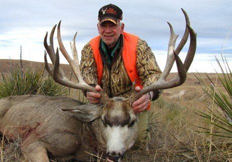 Man in camouflage and orange vest holding large deer antlers in a grassy field.