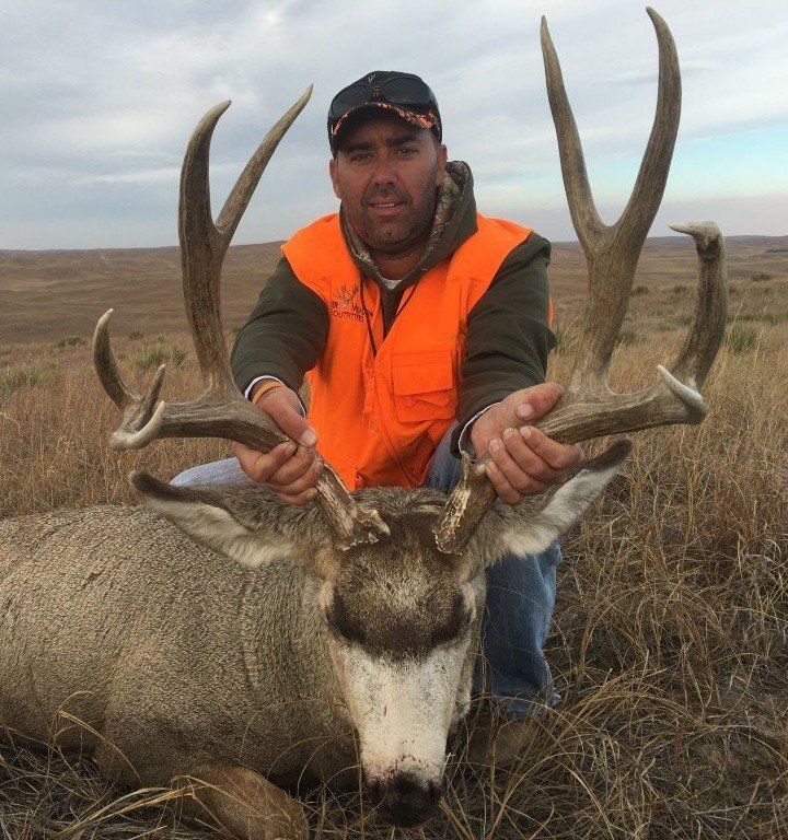 Man kneeling with a large buck he harvested, in an open field. Man wears orange vest and cap.