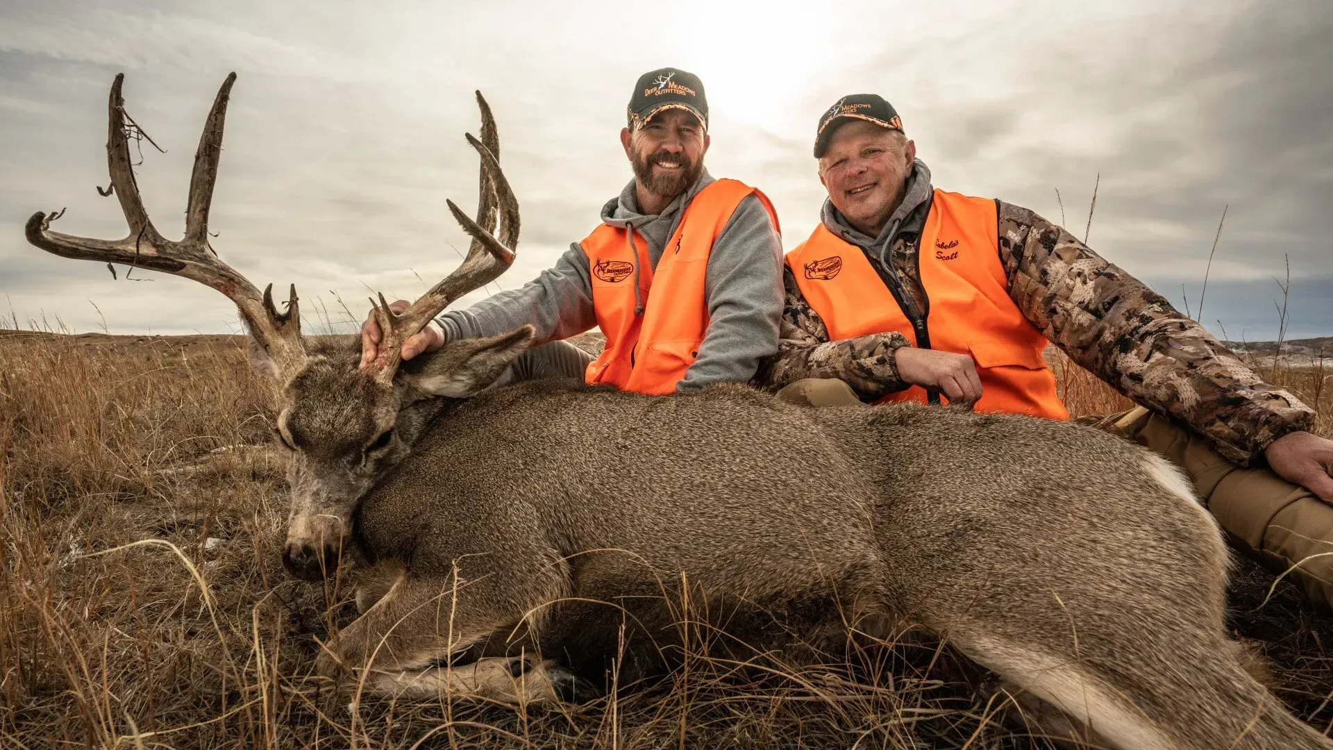 Two men in orange vests pose with a large buck in a field.