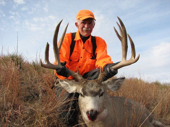 Hunter in orange shirt and cap with a large buck; outdoors.