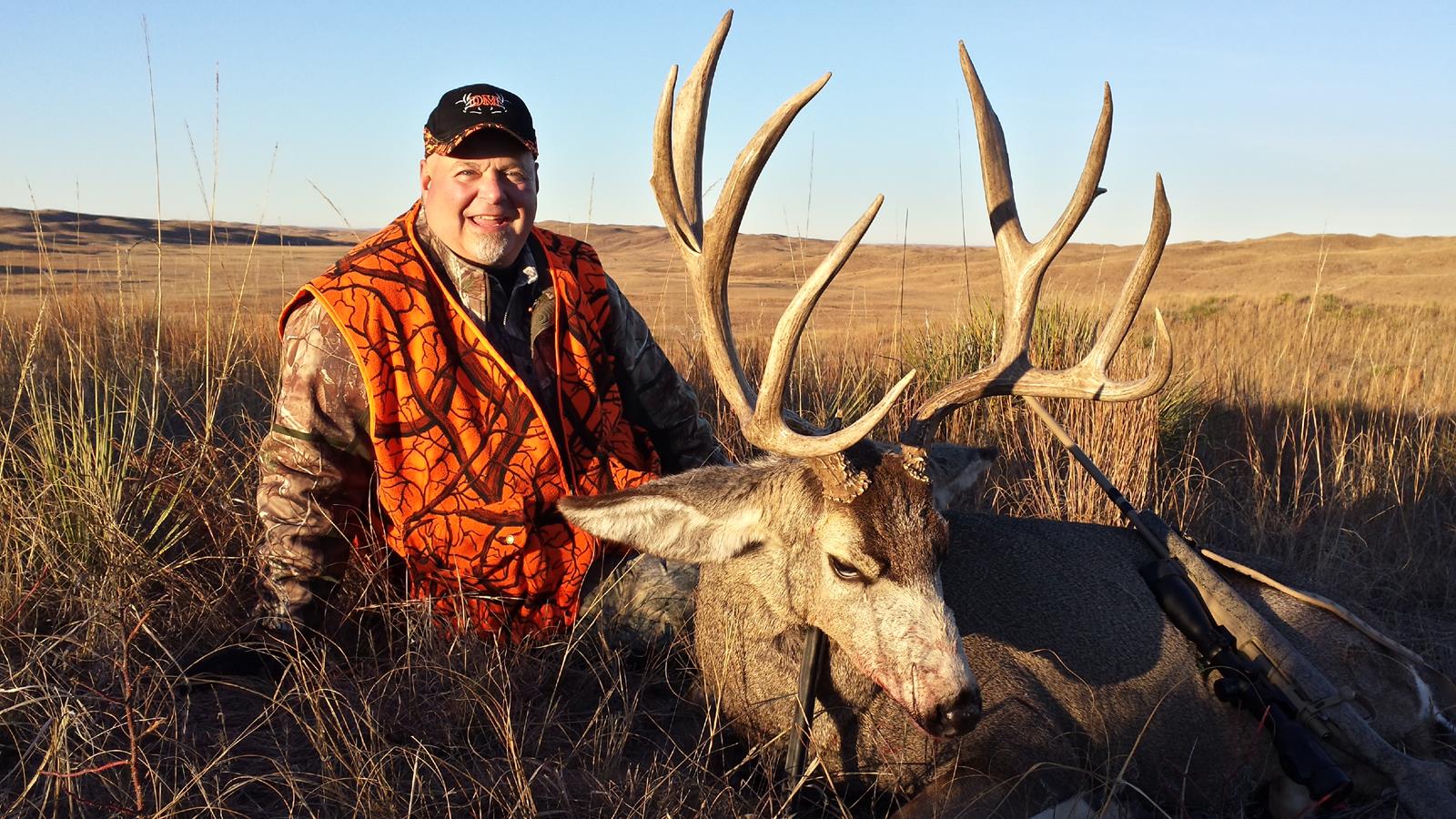 Hunter in orange vest smiles next to a large-antlered deer in a field.