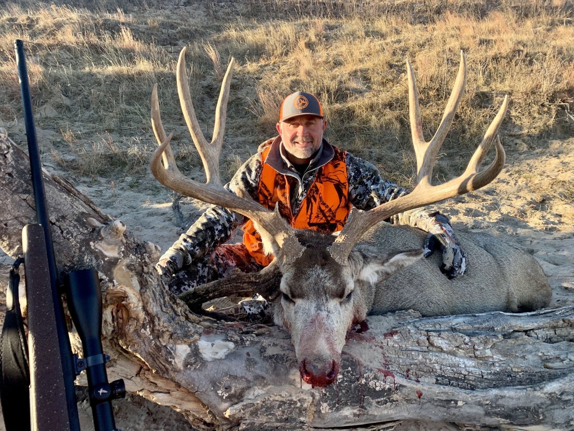 Man in orange vest smiles, posing with large dead deer, antlers prominent. Rifle in the foreground.