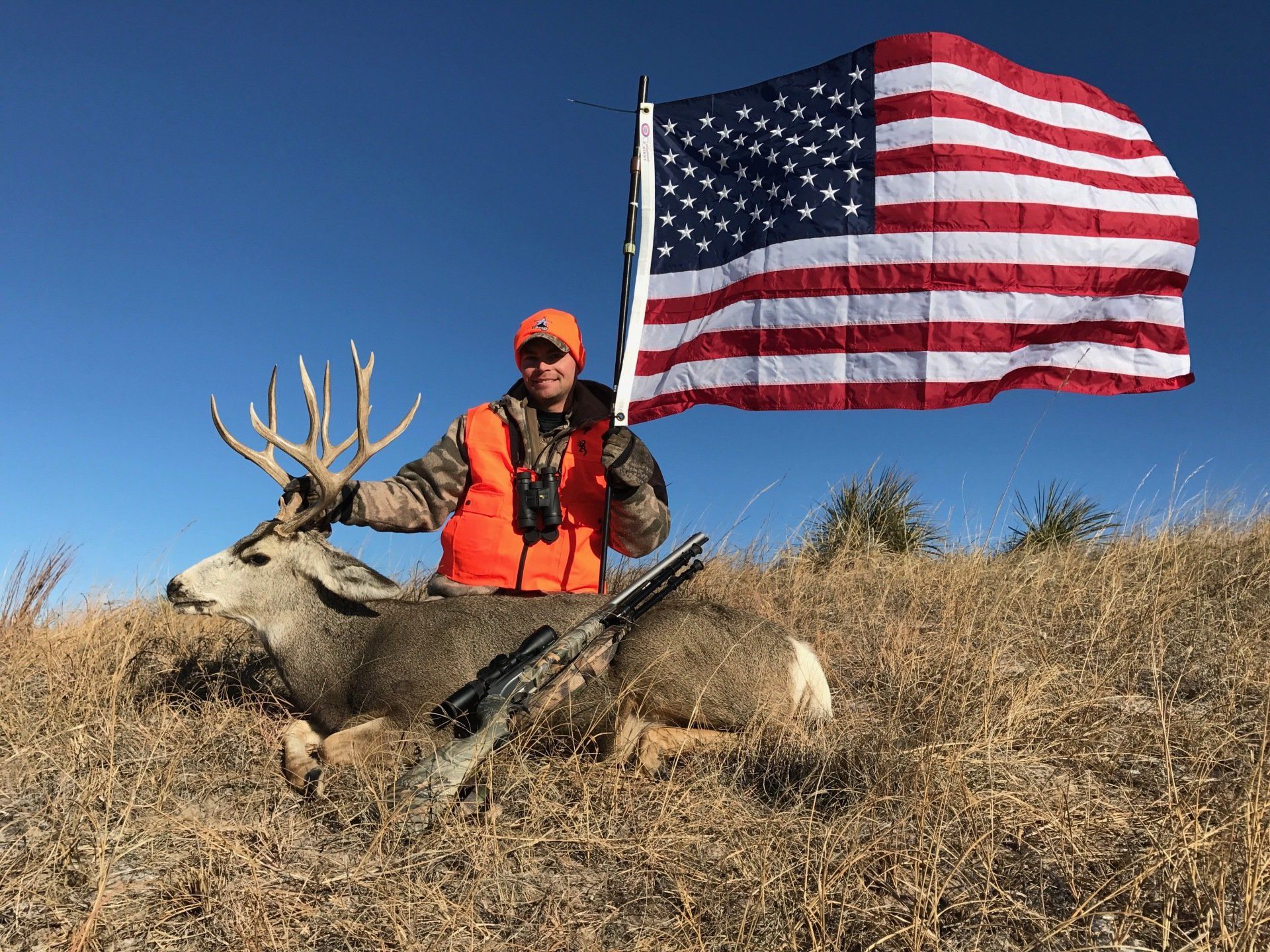 Hunter poses with large buck and American flag in field. Man wears orange vest and hat, holding rifle.
