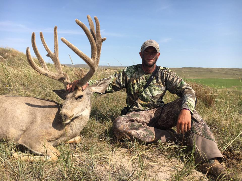 Hunter in camouflage kneels beside a large buck with impressive antlers in a grassy field.