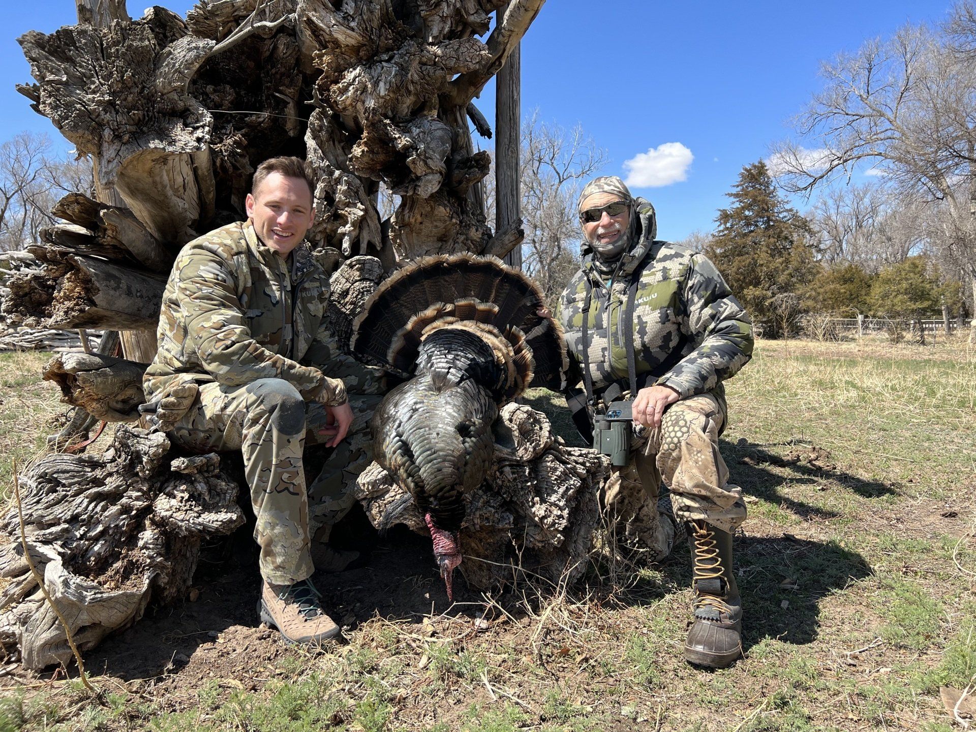 Two men in camo pose with a dead turkey in a field, smiling.