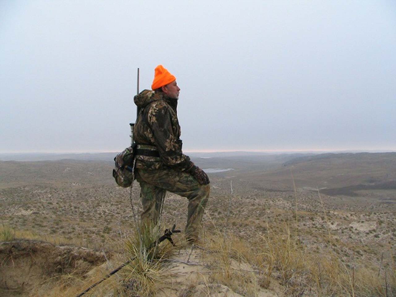 Hunter in camouflage with orange hat stands on a cliff, overlooking a vast landscape under a cloudy sky.