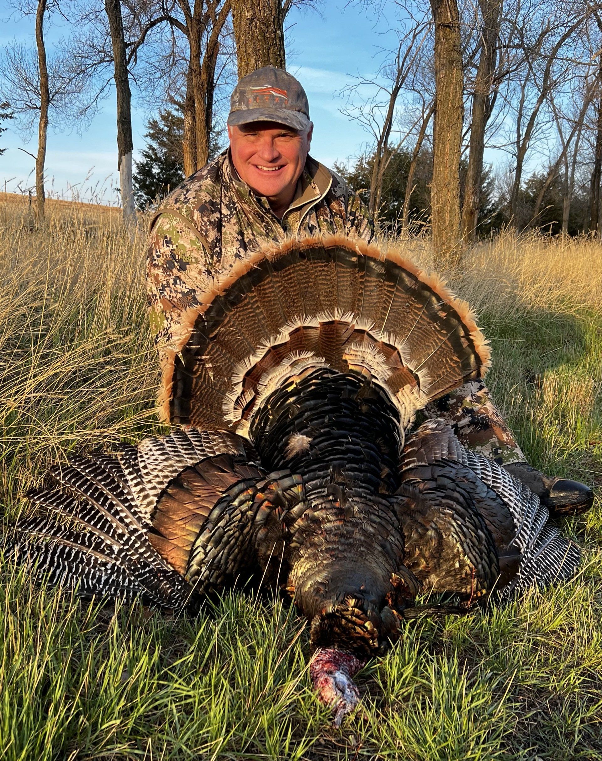 Man in camouflage proudly displays a large turkey with spread feathers in a grassy field.