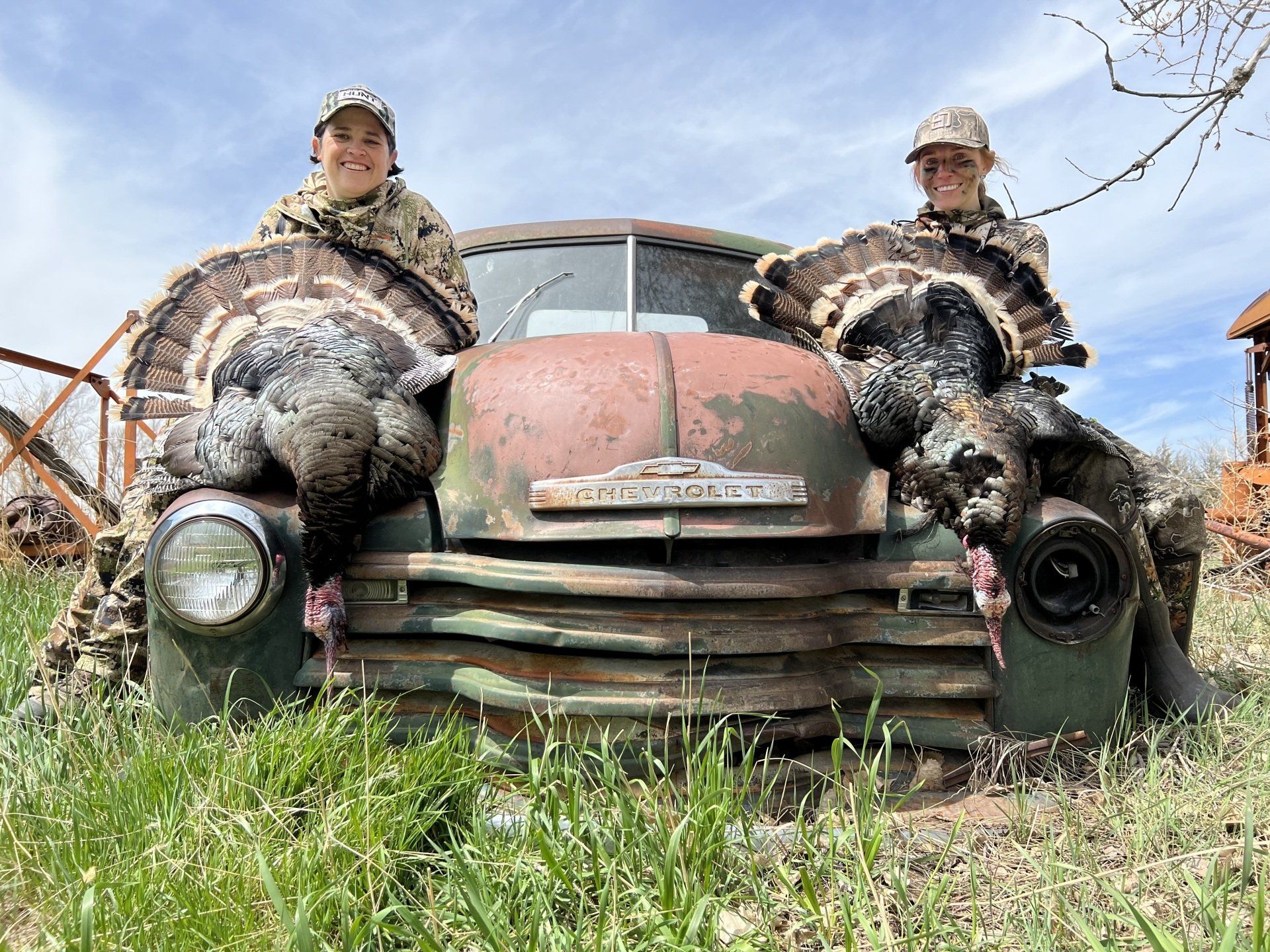 Two people posing with wild turkeys on an old Chevy truck. Camouflage clothing, green grass, outdoors.