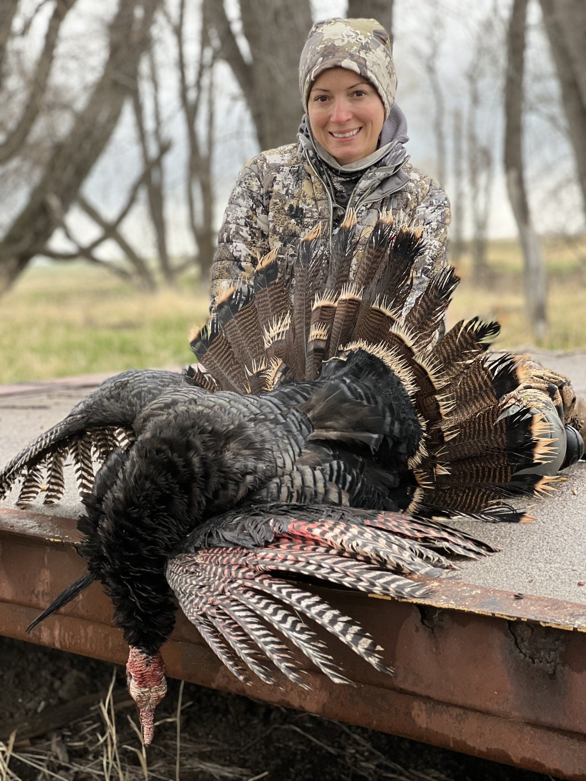 Woman in camo with a harvested turkey; outdoors, smiling.