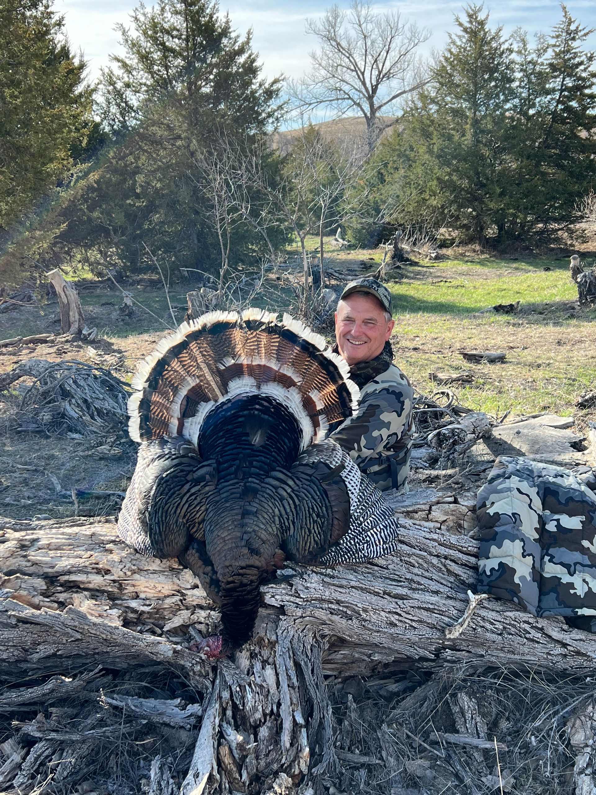 Man in camo holding a wild turkey with a fanned tail, posing outside in a wooded area.