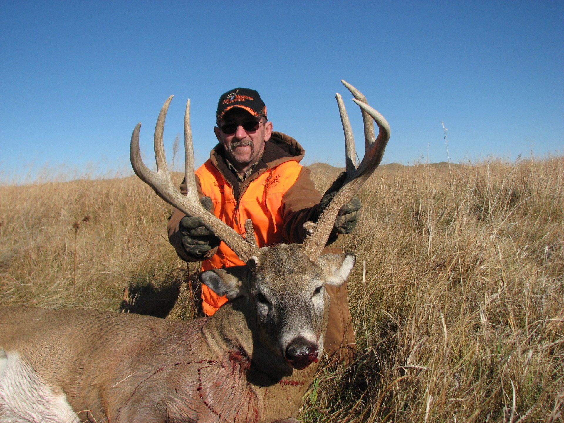 Hunter in orange vest kneeling with a large-antlered buck in a grassy field.