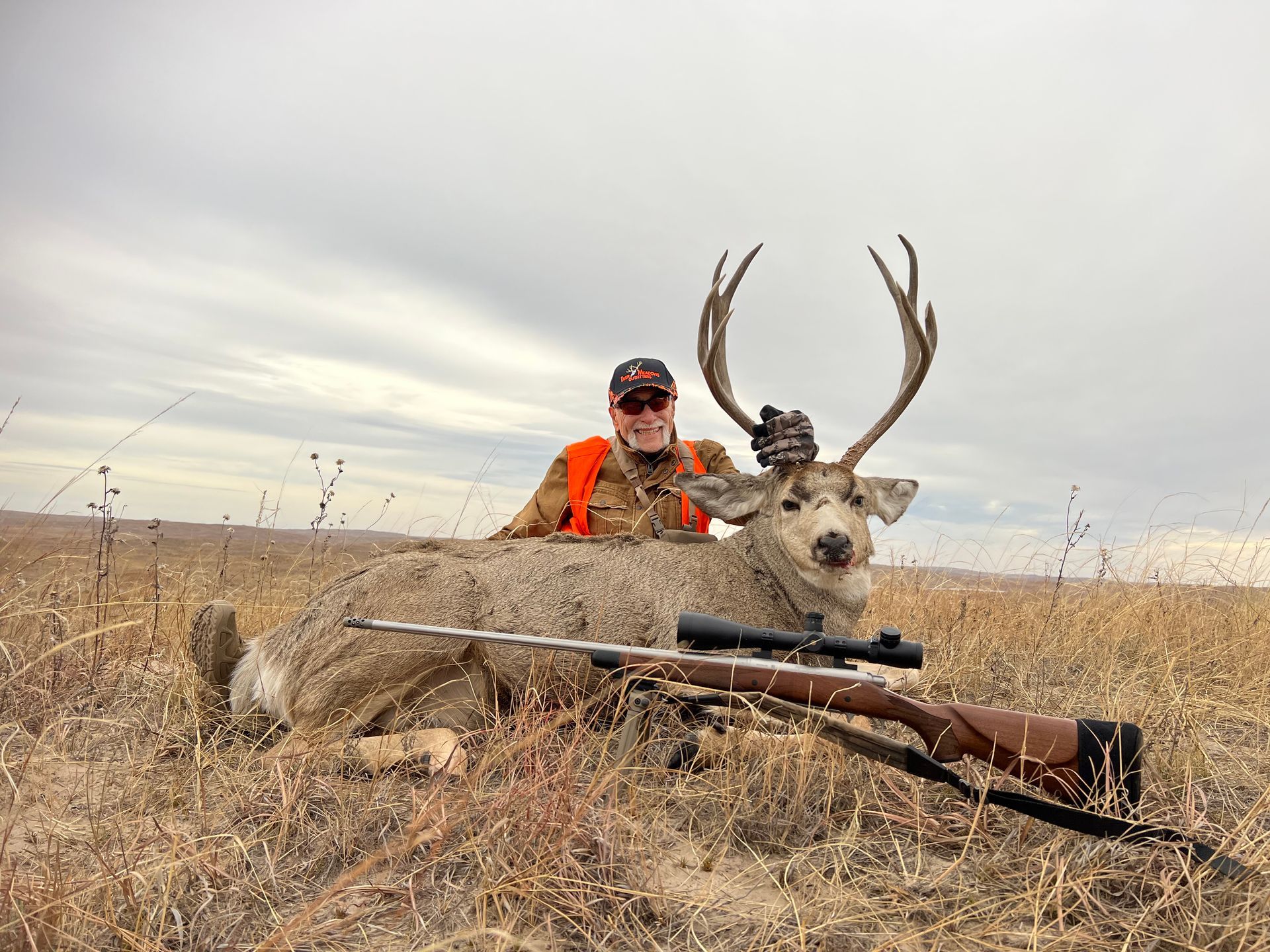 Hunter in orange vest poses with a large buck and rifle in a field. Overcast sky.