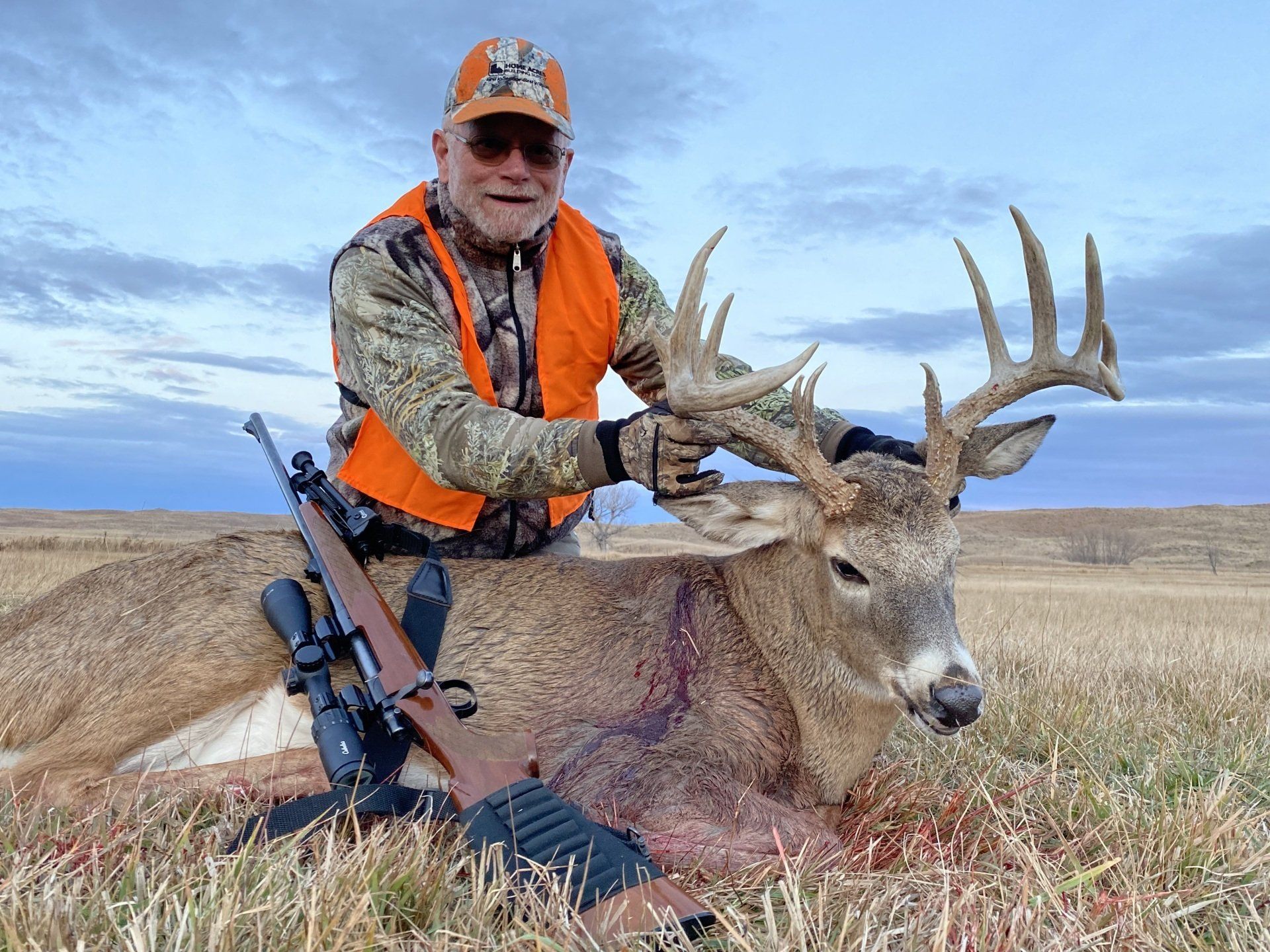 Hunter with a large buck he harvested in a field; hunter smiling, wearing orange vest.