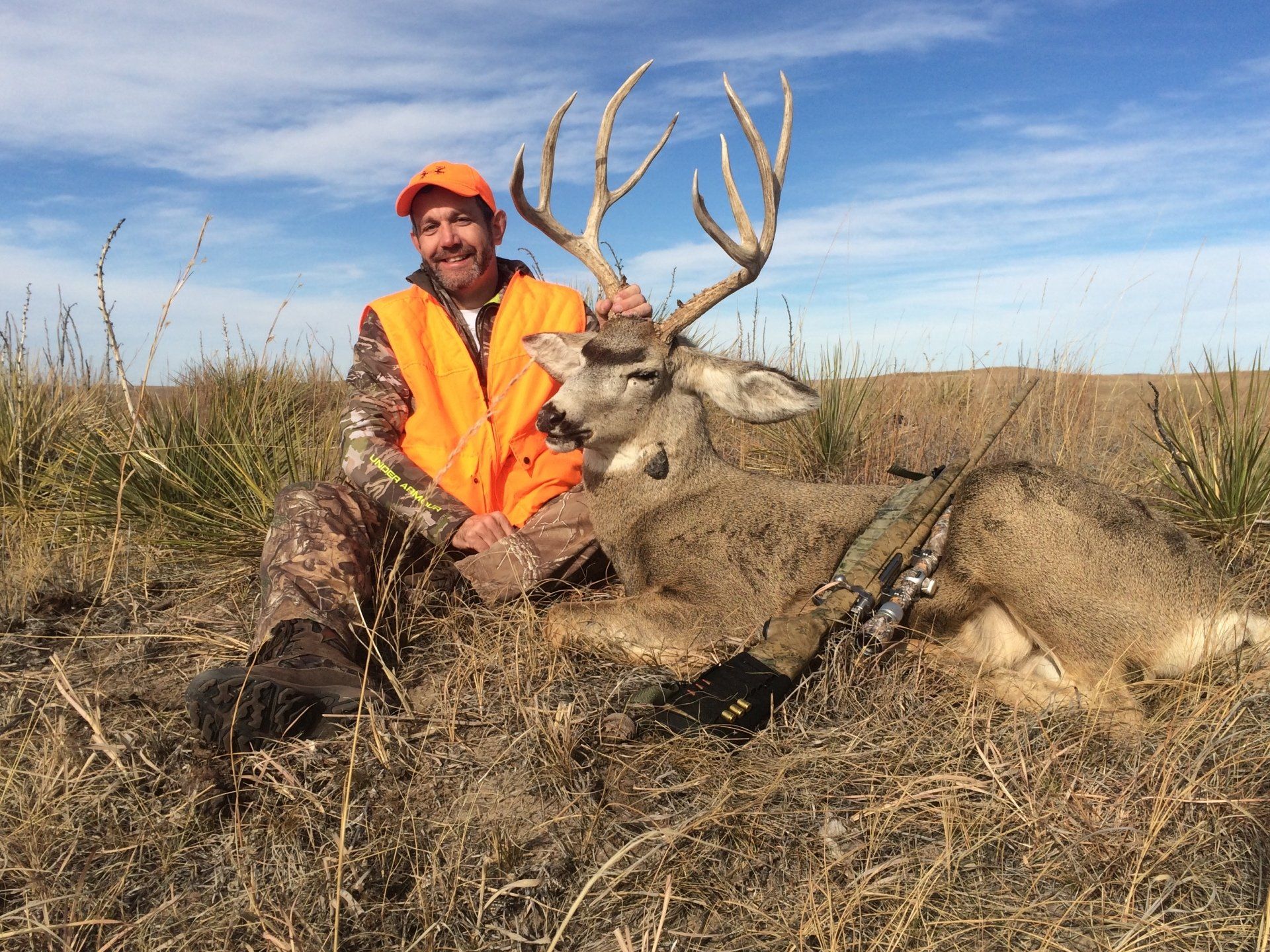 Hunter kneels beside a large deer he harvested. He wears orange vest in a prairie setting.