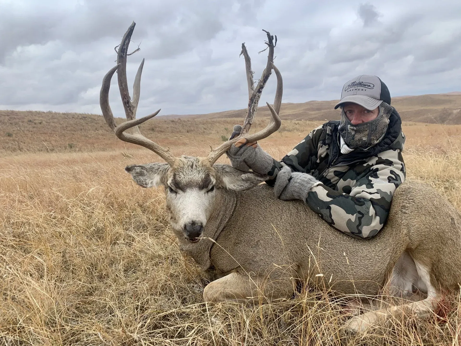 Hunter sits on a large buck deer in a field, holding its antlers. Overcast sky.