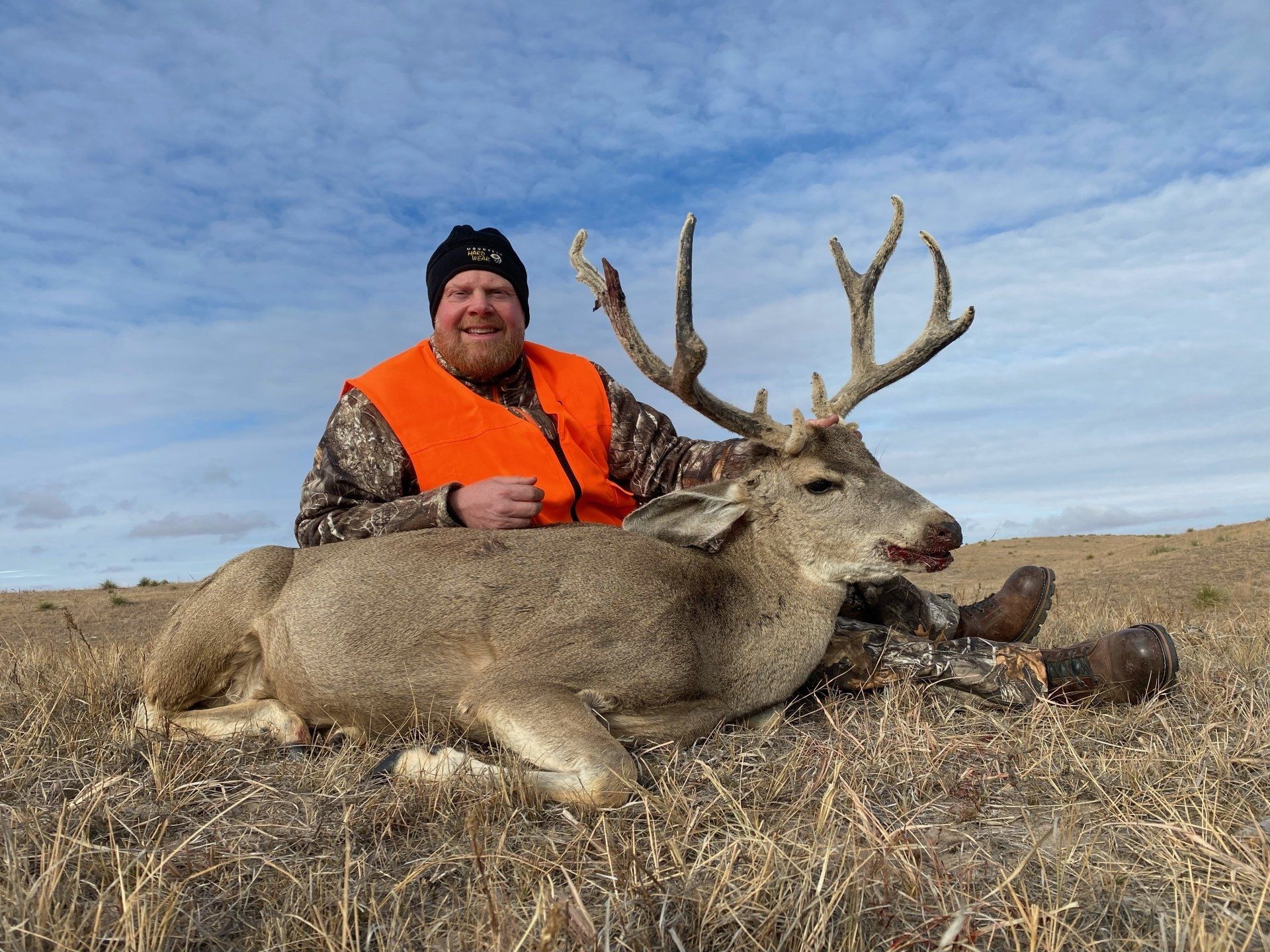 Man in orange vest sits next to a harvested buck with large antlers in a grassy field.