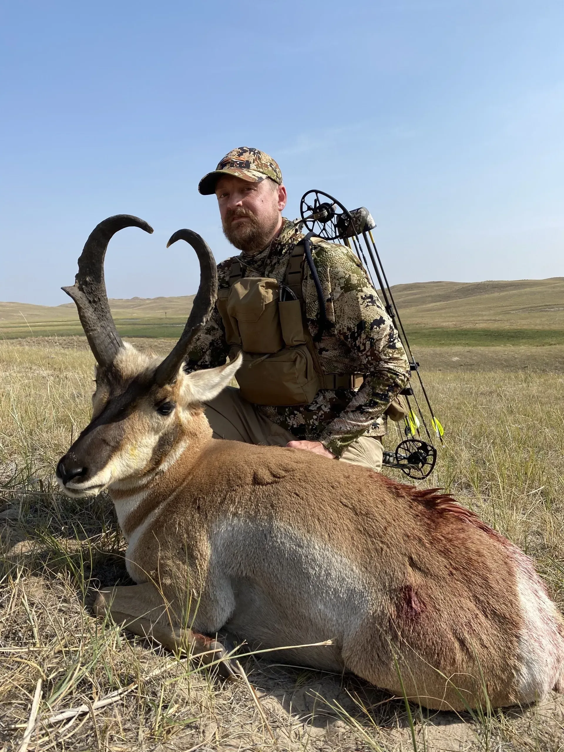 Man in camo kneels next to a harvested pronghorn in a grassy field.