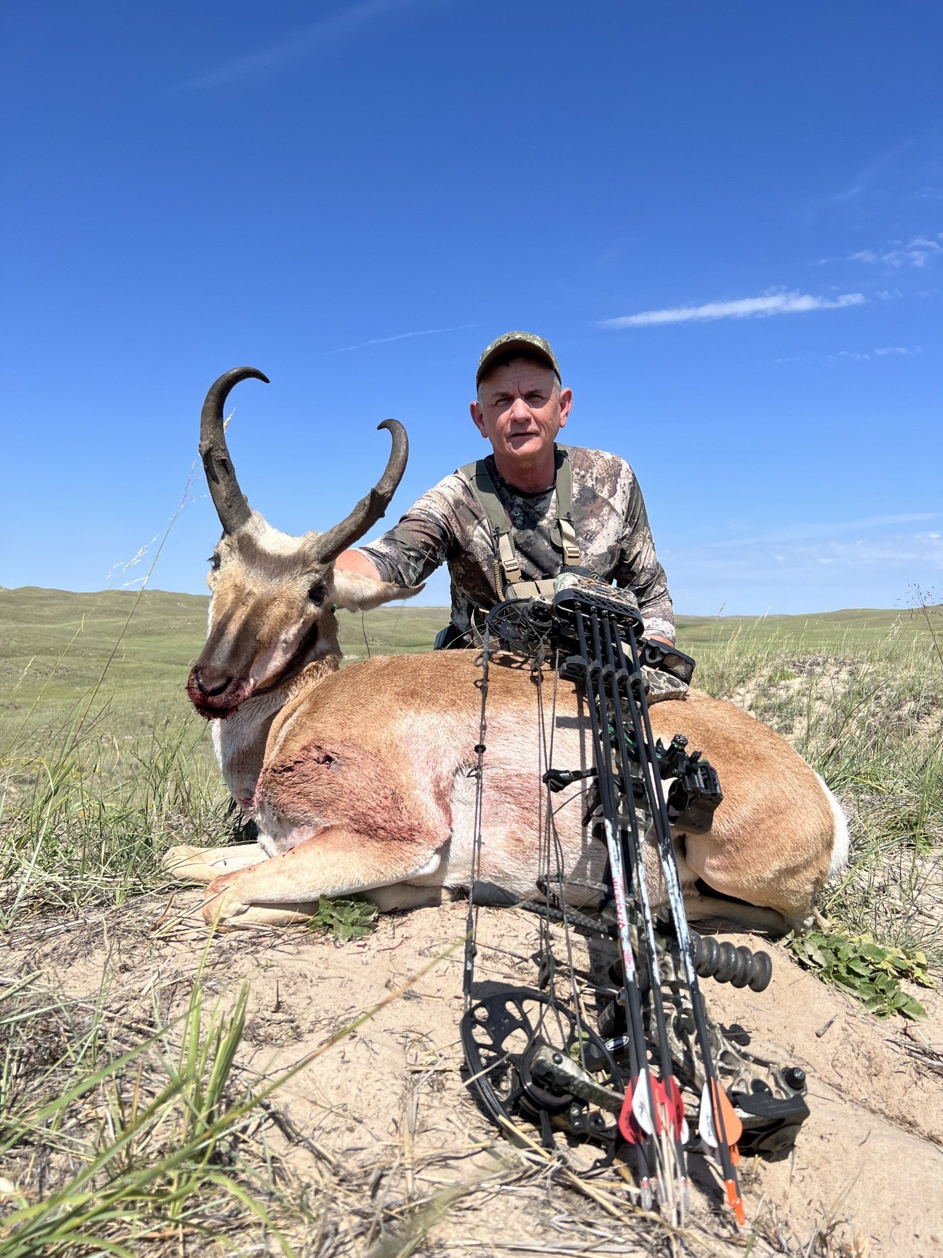 Hunter with a pronghorn antelope and bow. Tan fur, blue sky background. Man smiling, outdoors.