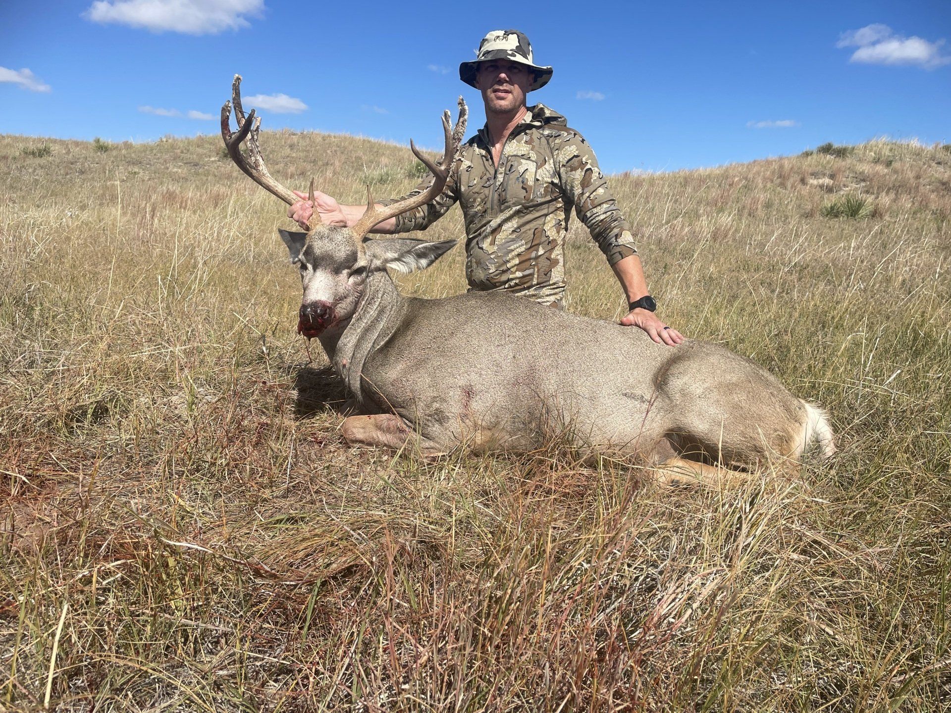 Man in camo with a large buck in a grassy field; the buck has large antlers.