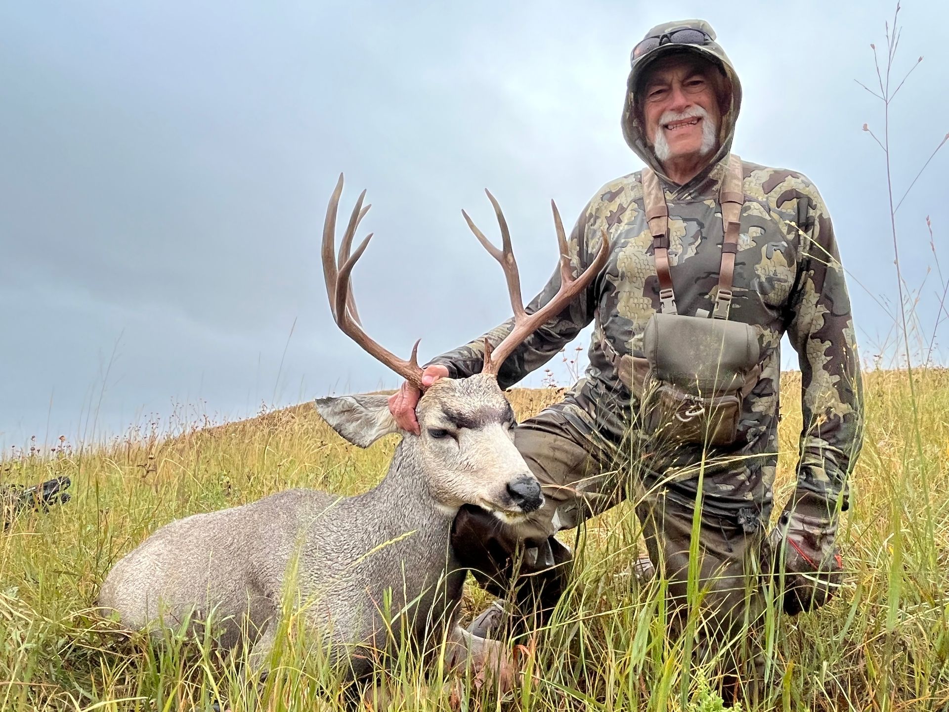 Man in camo kneels beside a harvested deer in tall grass. Cloudy sky.