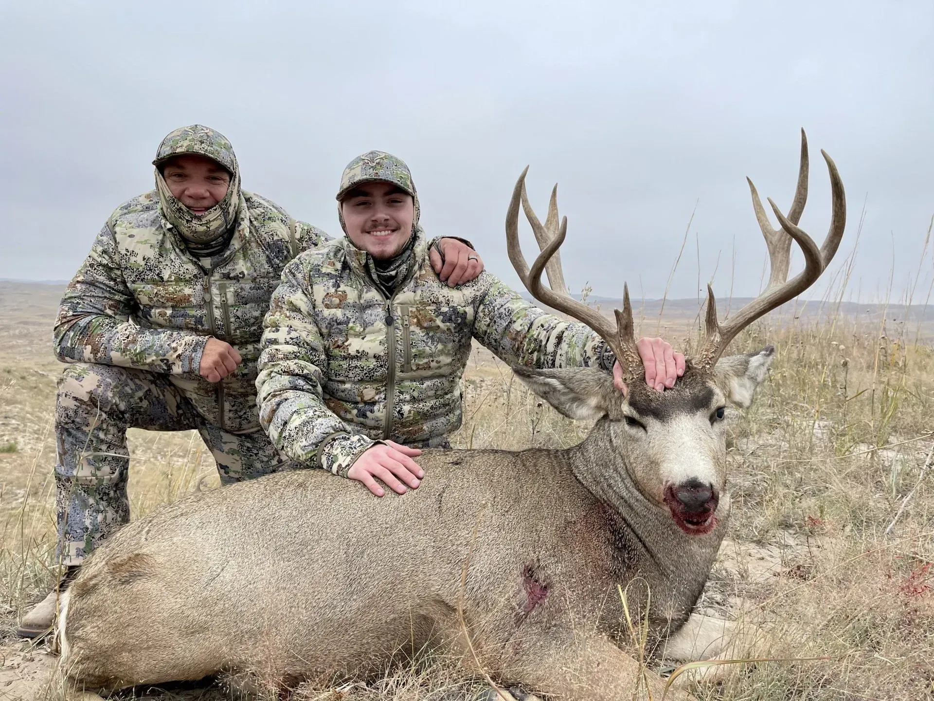 Two men in camouflage pose with a large, harvested buck in a field.
