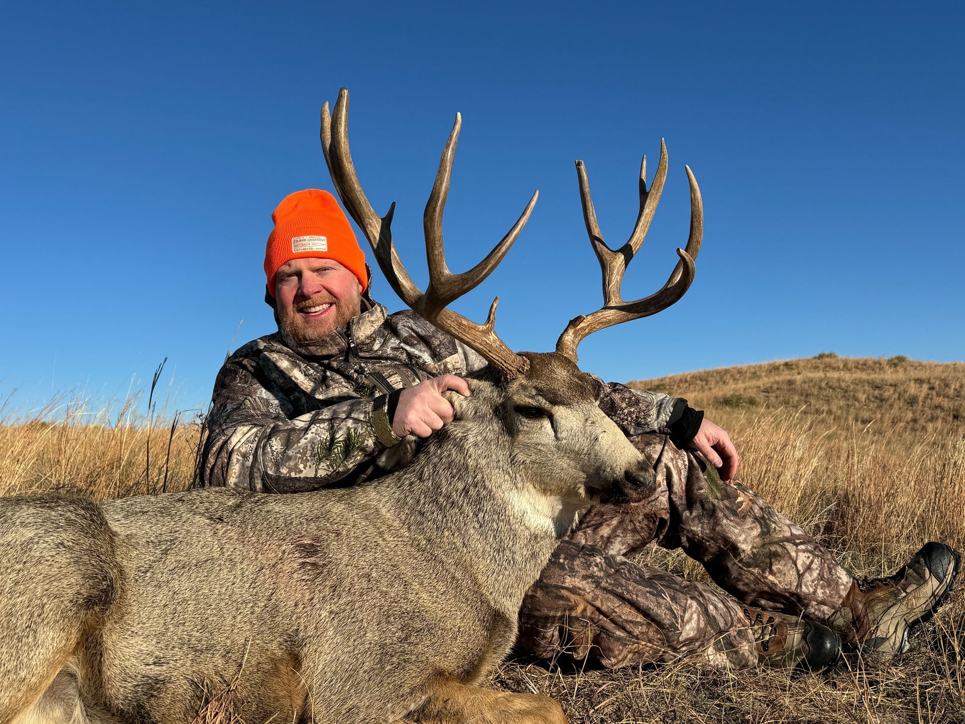 Man in camo and orange hat smiles, posing with a large buck in a field under blue sky.