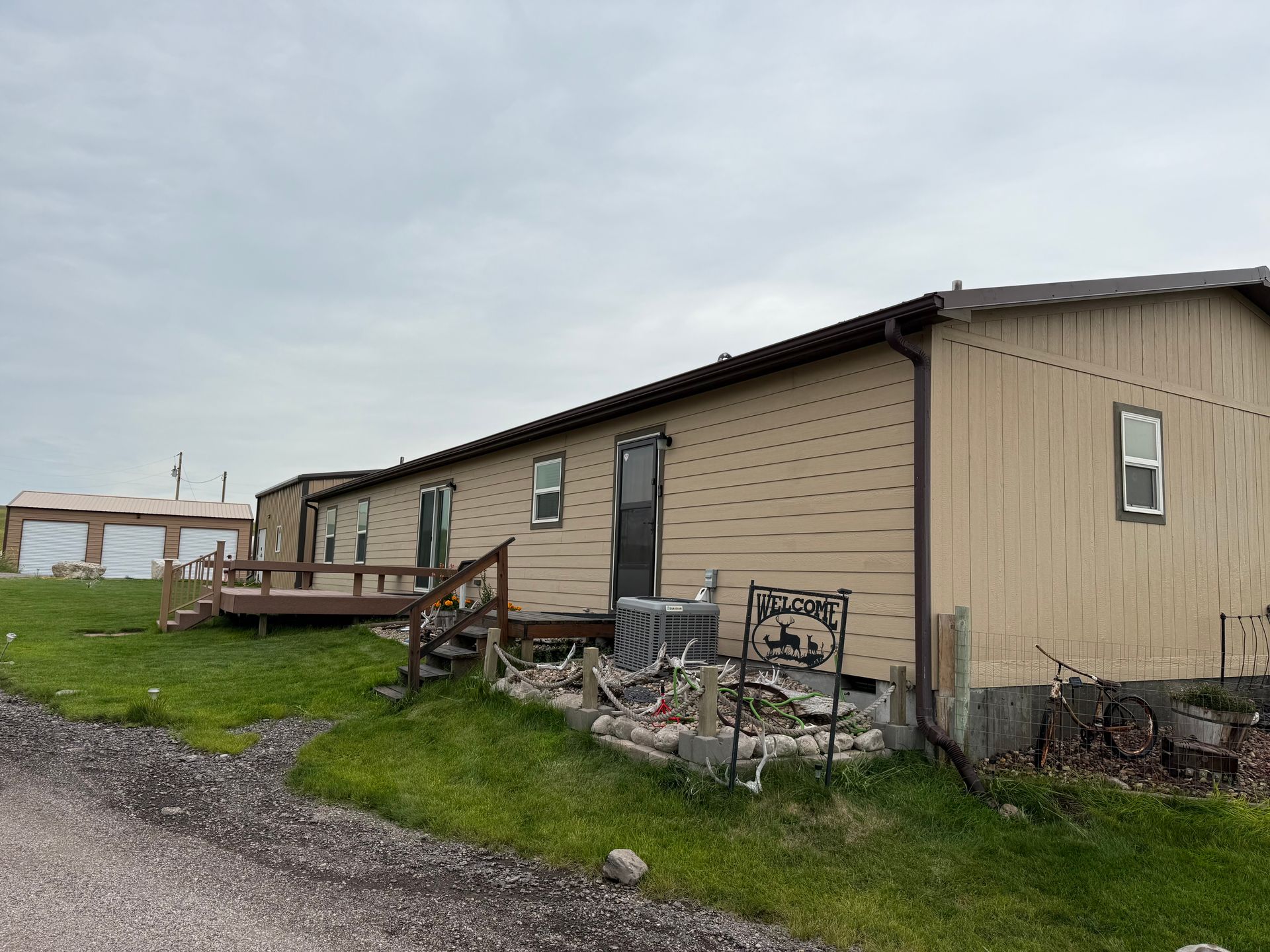 Tan mobile home with a deck, on a grassy hill. Cloudy sky overhead.