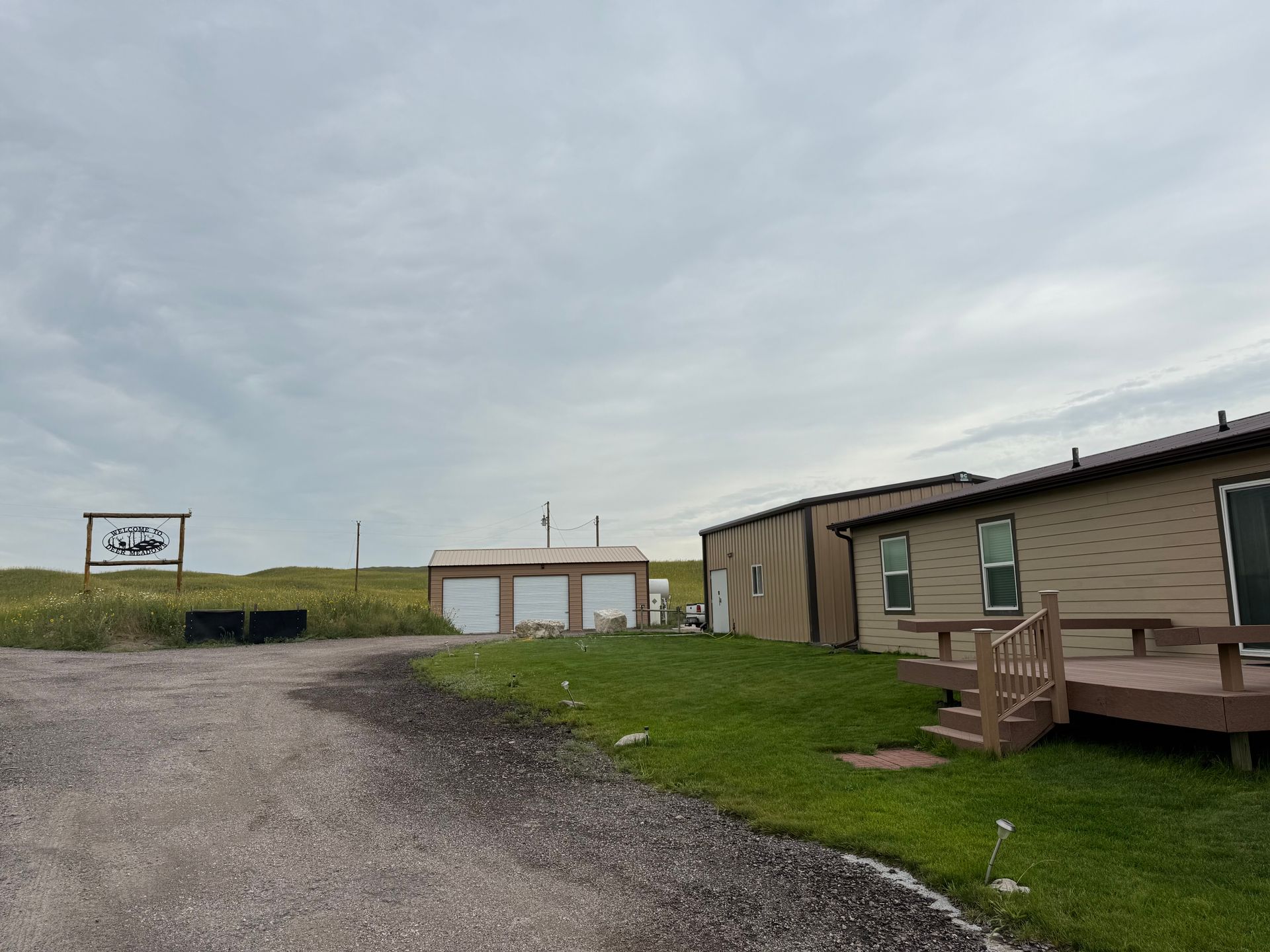 Gravel driveway leads to rural buildings on a cloudy day. Two-car garage and a house with a deck are visible.