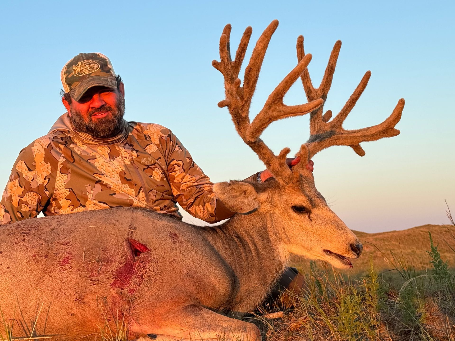 Man in camo with large-antlered deer he harvested in field at sunset.