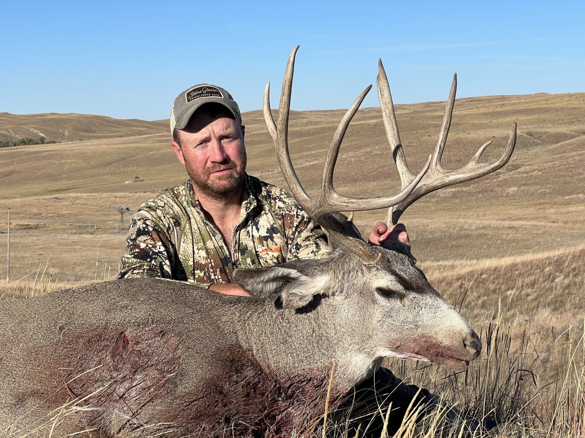 Man in camo with a large buck he has harvested in a grassy field under a blue sky.