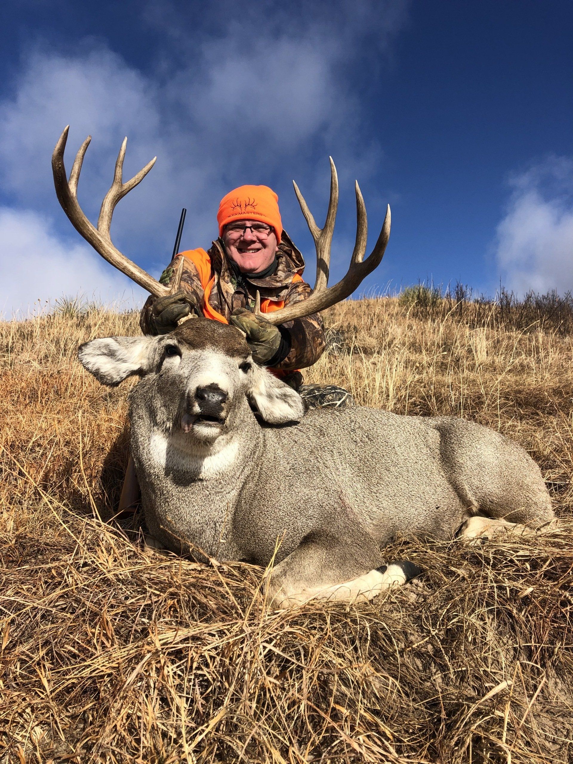 Hunter in orange hat smiles with a large buck, antlers up, on a hillside under blue sky.