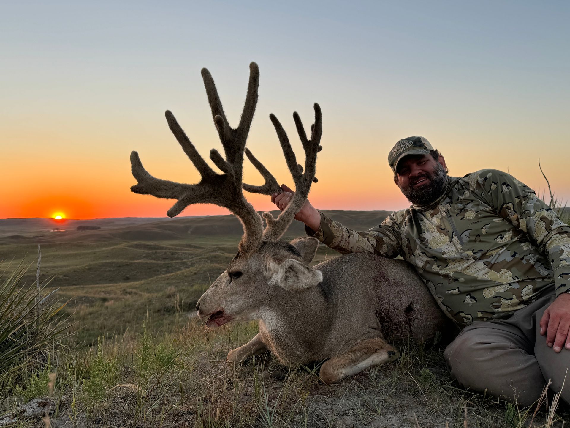 Hunter with a large buck, posing in a field at sunset.