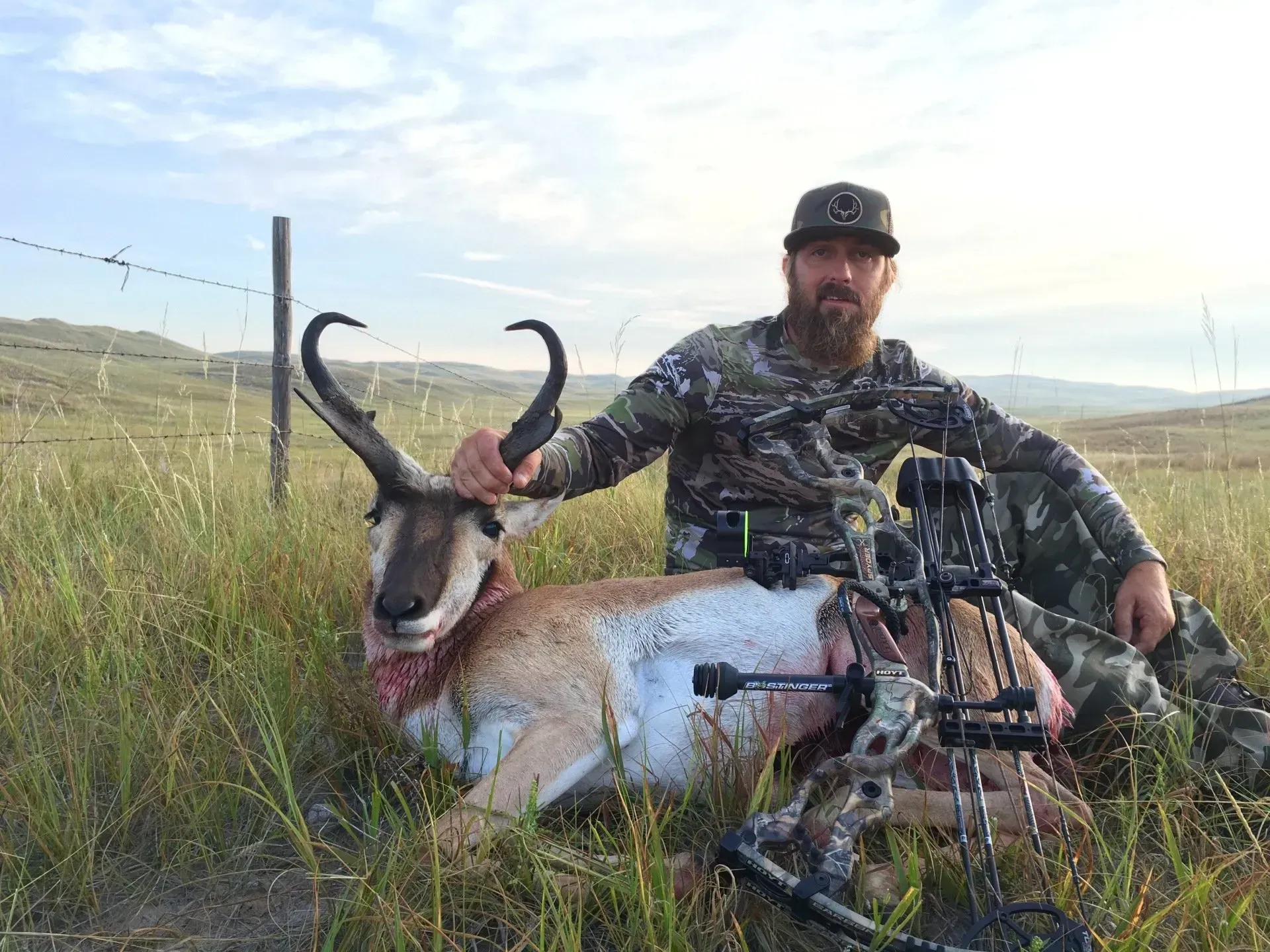 Hunter with a bow sits next to a harvested pronghorn antelope in a grassy field.