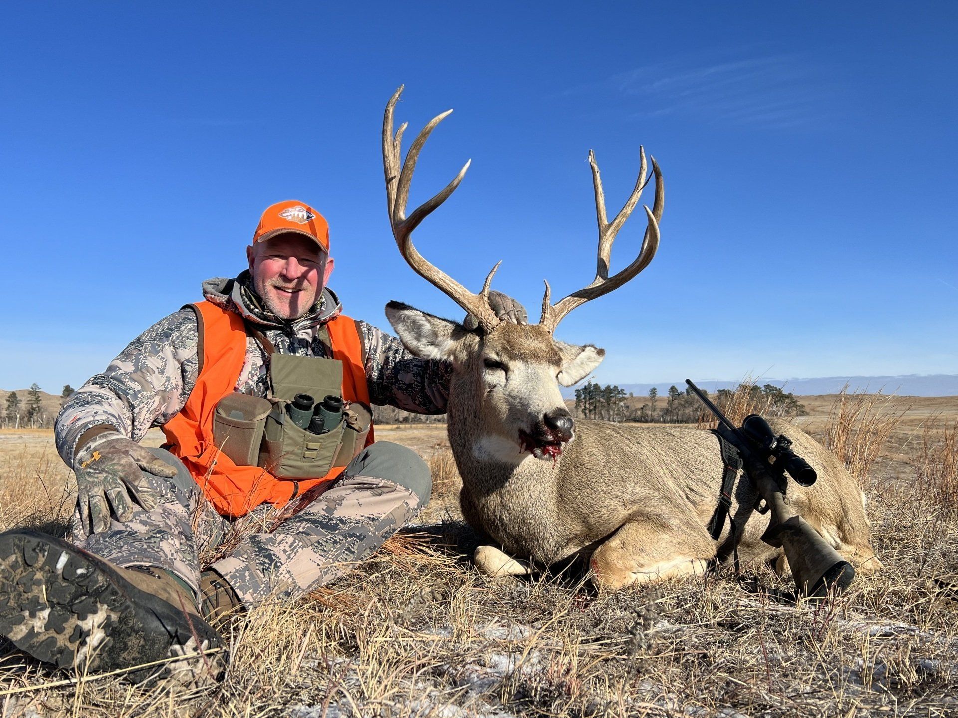 Hunter kneels beside a large mule deer buck with antlers. Wearing orange vest, he smiles in a field on a sunny day.