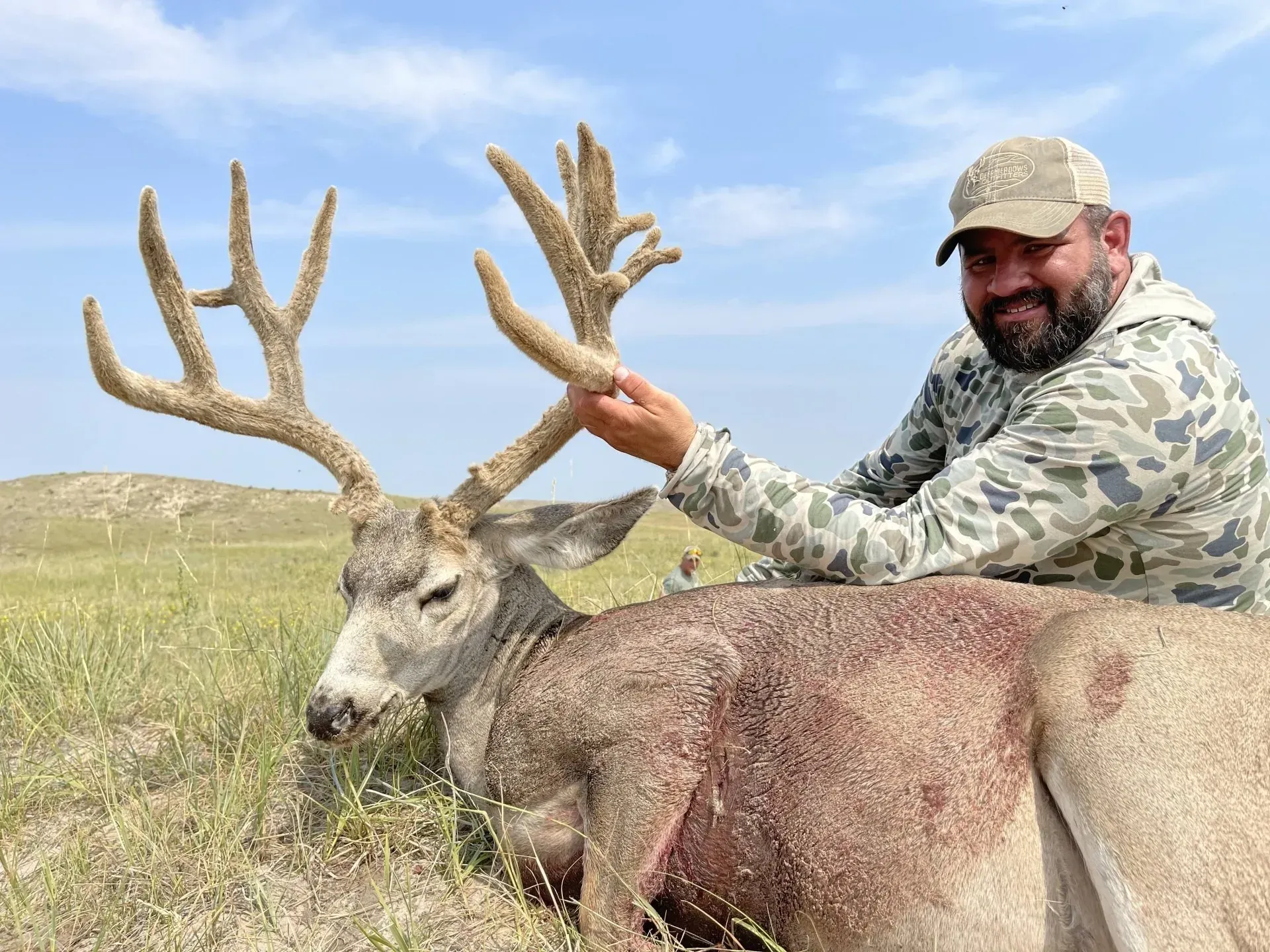 Man in camo with a large buck he harvested, in a grassy field, smiling, under a blue sky.