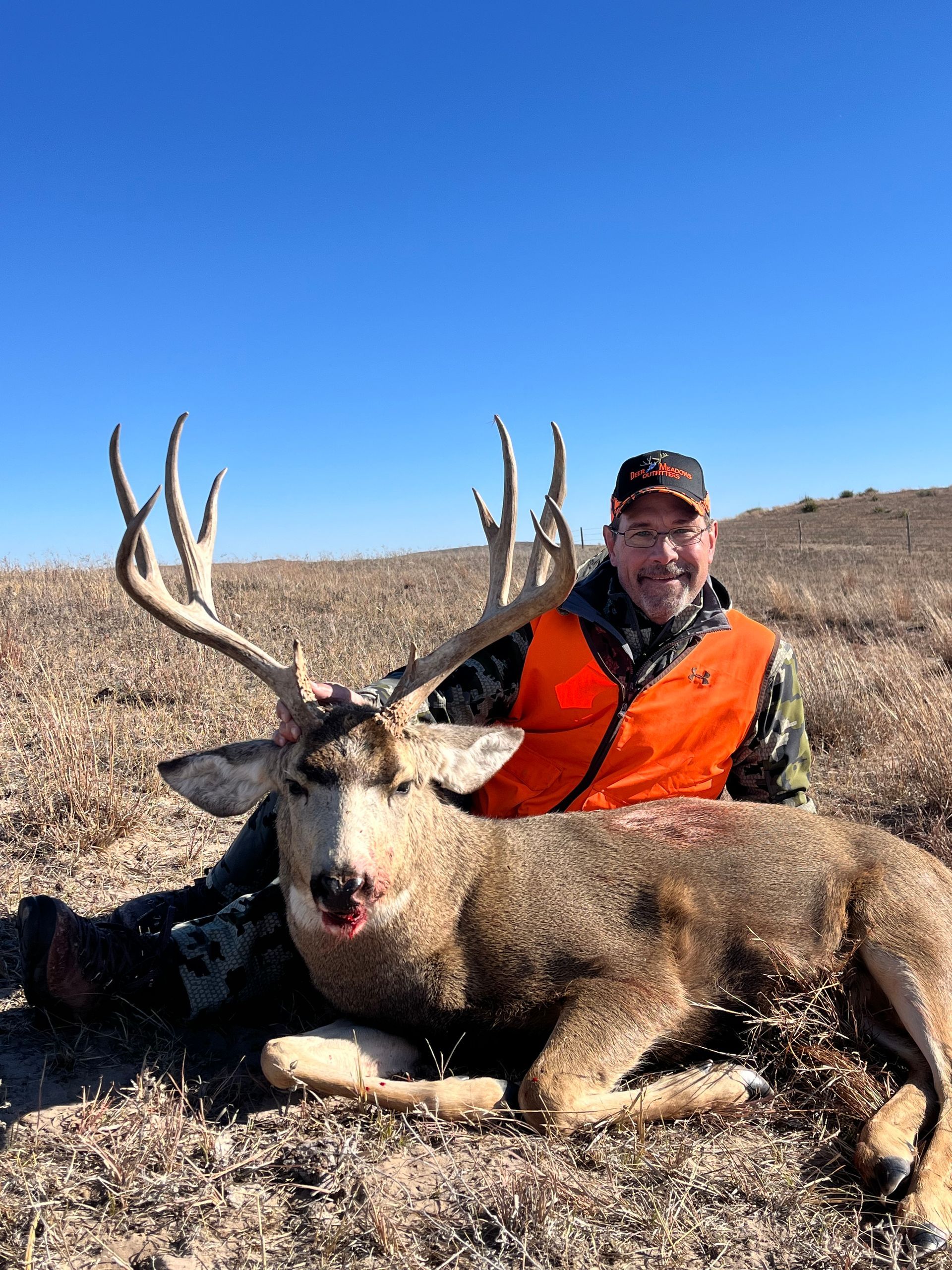 Man in orange vest kneels with a large mule deer buck, smiling. Outdoors, blue sky.