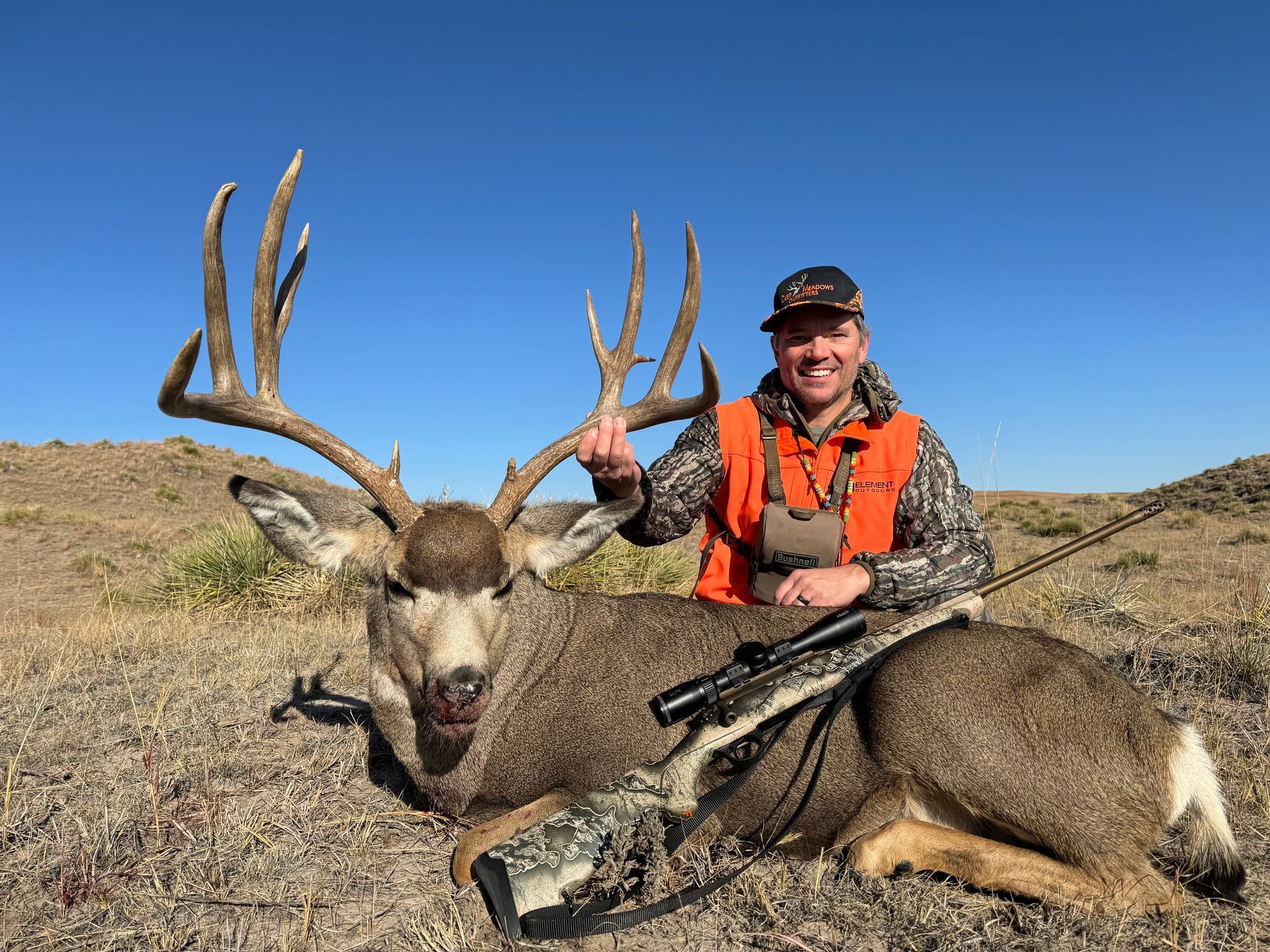 Hunter smiles with a large buck. Deer lies on the ground, hunter holds its antlers, rifle beside them, clear blue sky.